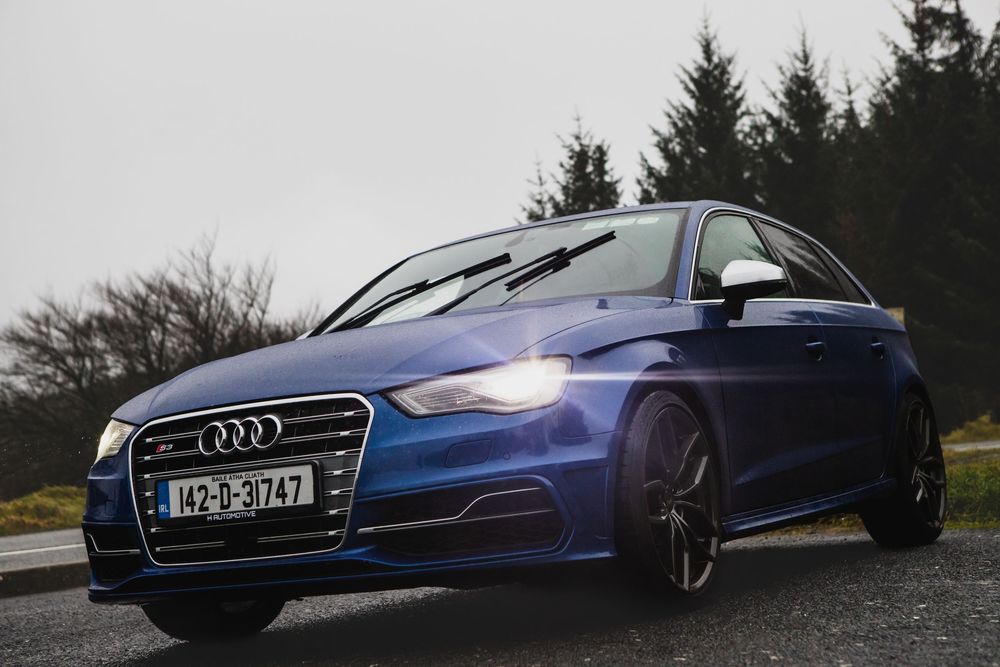Blue Audi S3 sedan with Irish license plate parked on a road with headlights on.