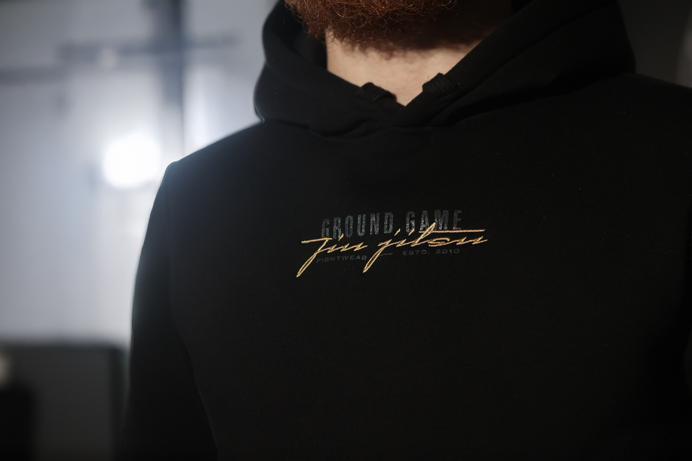 Man wearing black Ground Game hoodie with gold cursive logo embroidery on chest.