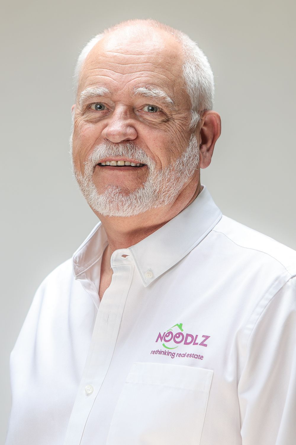 Smiling senior man with white hair and beard wearing a Noodlz Rethinking Real Estate branded white polo shirt.