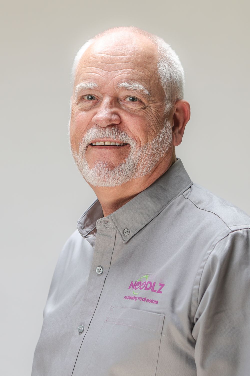Smiling older man with white hair and goatee wearing a gray Noodlz branded button-up shirt against a light background.