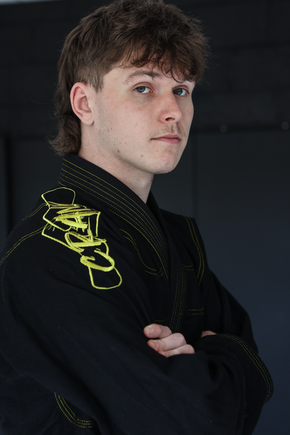 Young man in black BJJ gi with yellow embroidery standing with arms crossed.