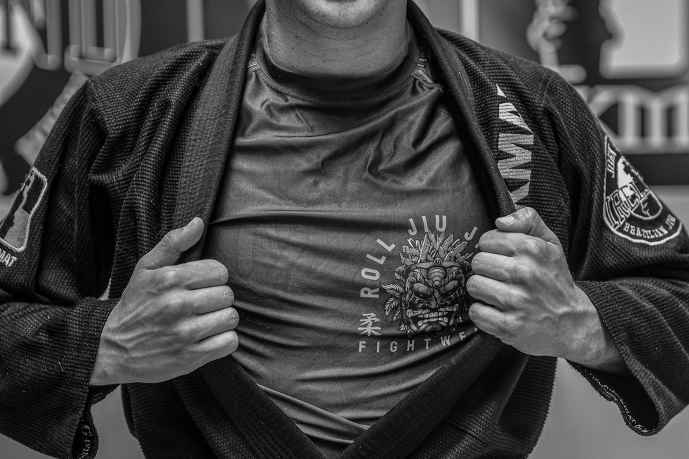 Athlete in black gi reveals Roll Jiu Jitsu branded rashguard underneath in black and white photo.