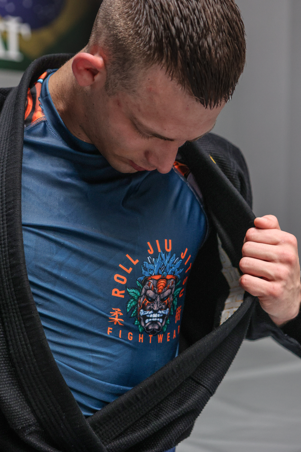 Male athlete in blue Roll Jiu Jitsu Fightware rashguard adjusting black gi jacket in gym.