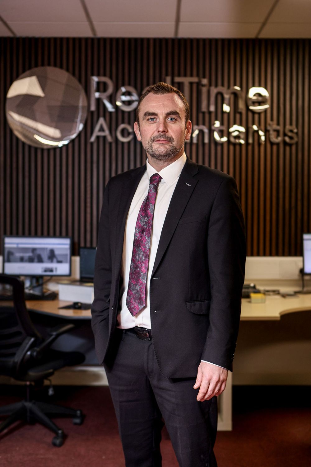 Professional man in dark suit standing before RealTime Accountants logo in modern office.