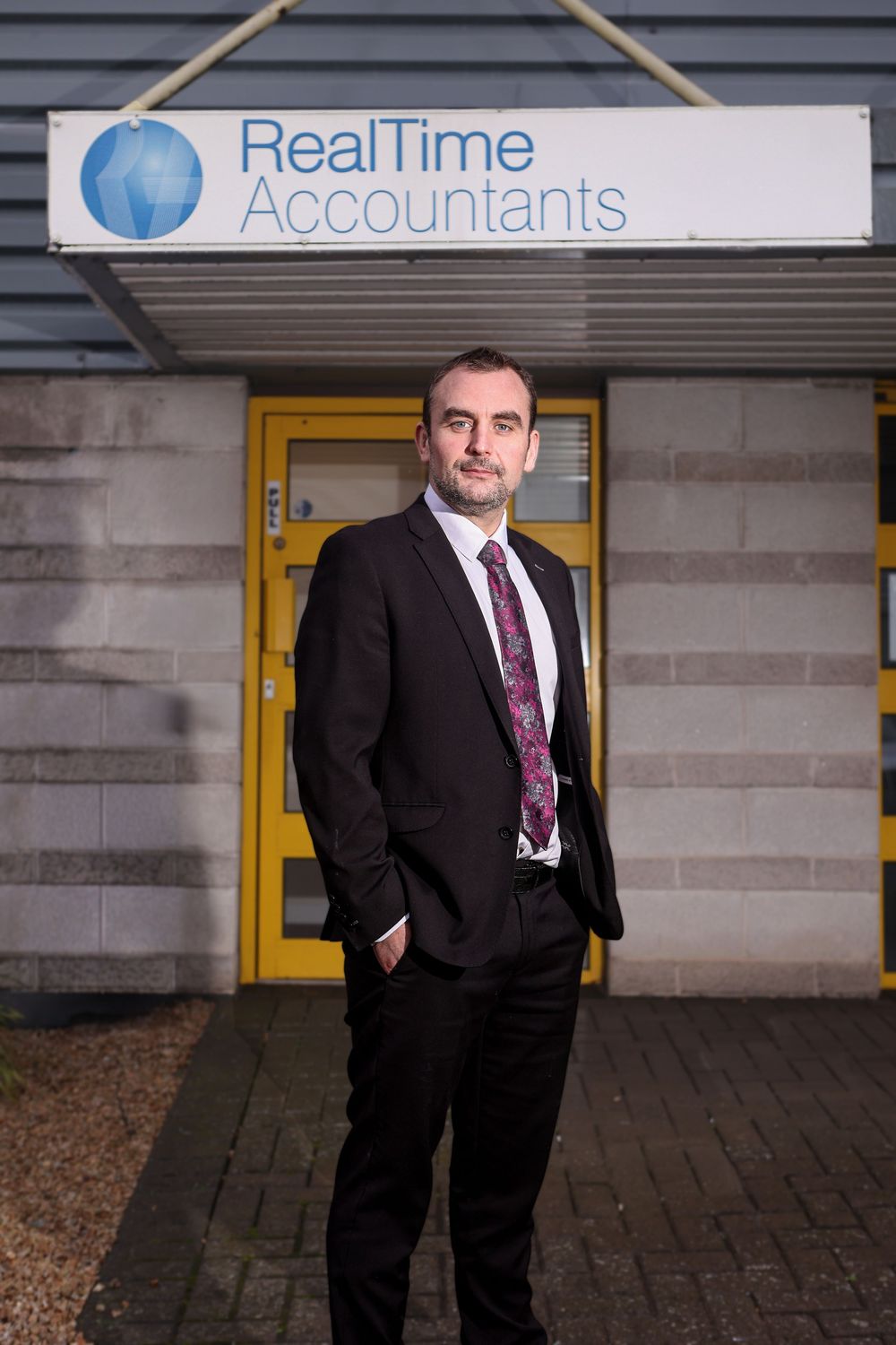 Businessman in dark suit standing outside RealTime Accountants office building entrance.