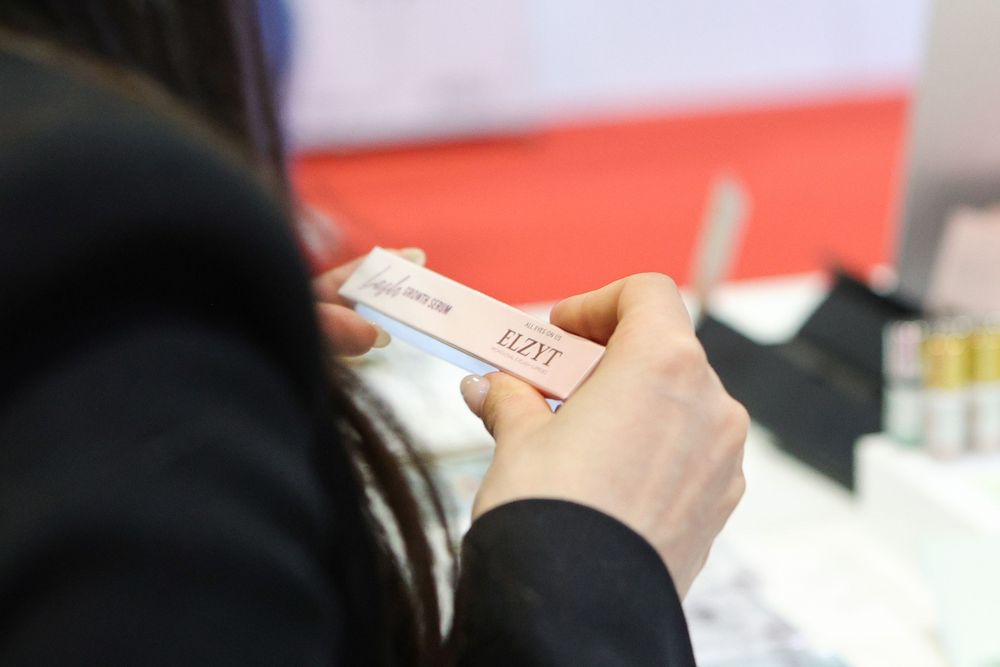Close-up of hands holding a nail file at a beauty trade show with cosmetic products in the background.