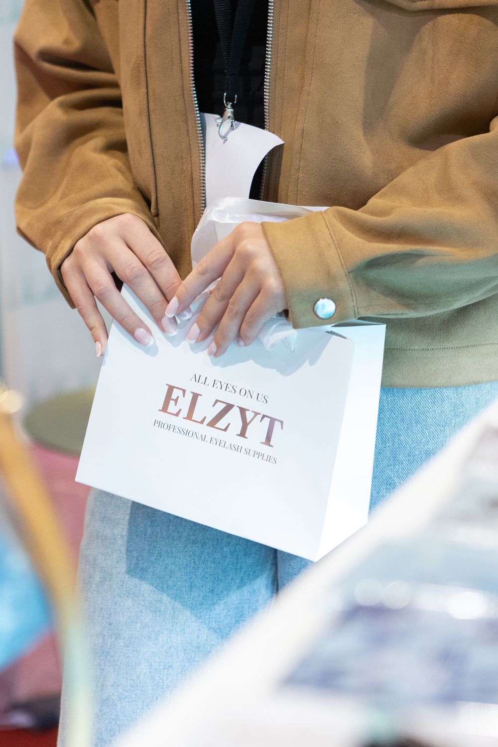Woman in brown jacket holding a white ELZYT professional eyelash studio shopping bag at a beauty event.