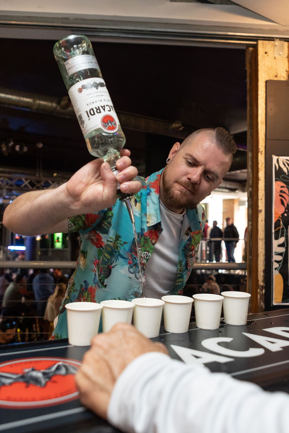 Bartender in floral shirt pouring Bacardi rum into paper cups at a bar event.
