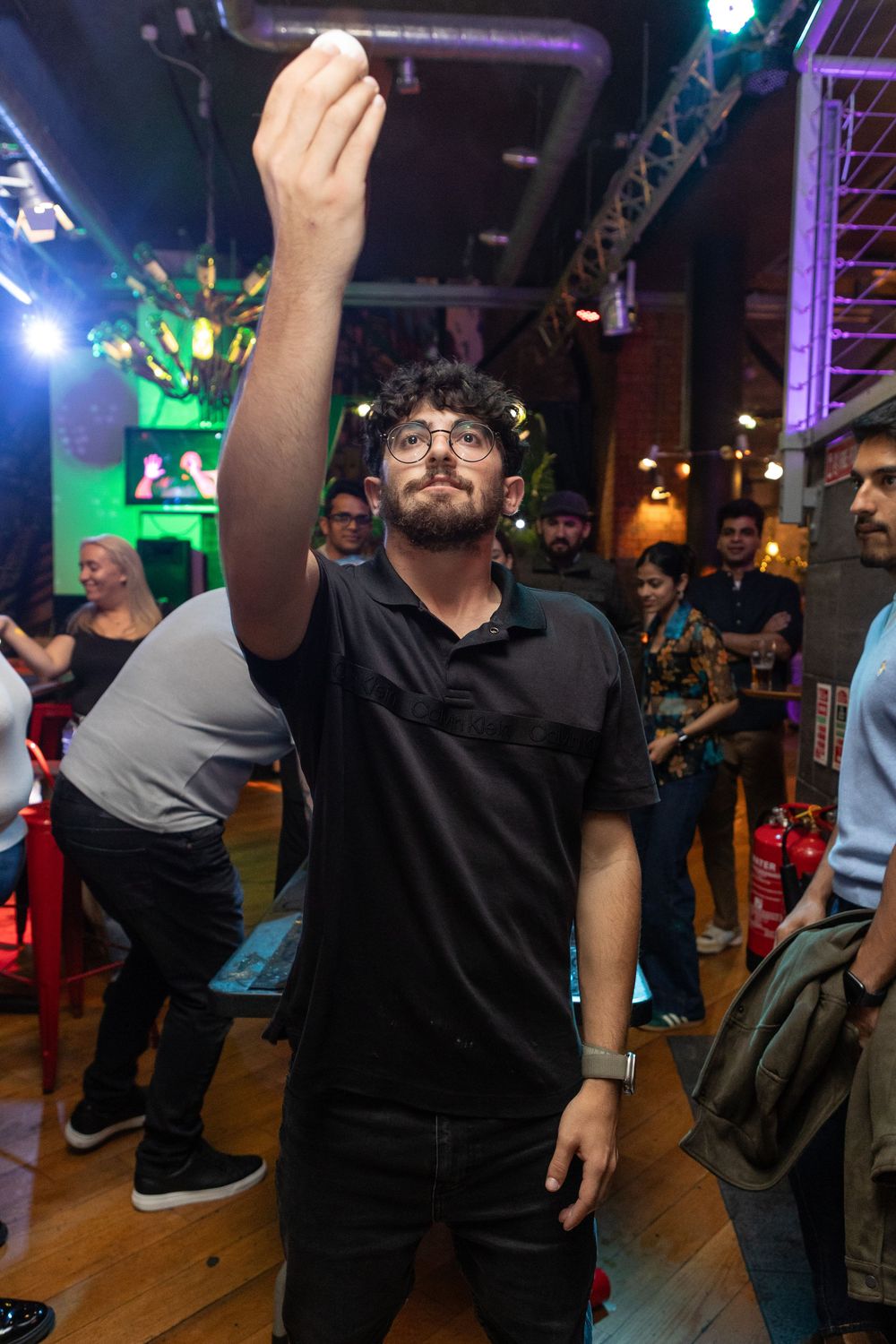 Bearded man with glasses raising hand in a lively indoor bar or event venue.