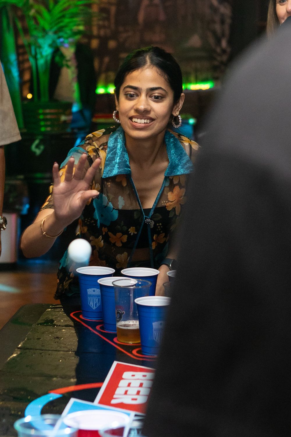 Smiling woman waving hand over beer pong table at a vibrant indoor social event.