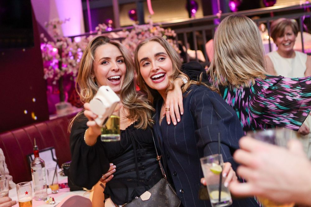 Two women smiling and taking a selfie at an elegant evening event, holding cocktails in a warmly lit venue.