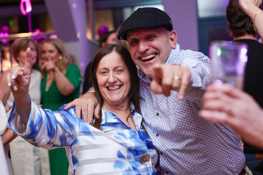 Two people smiling and posing together at a lively party event, with colorful lights in the background.