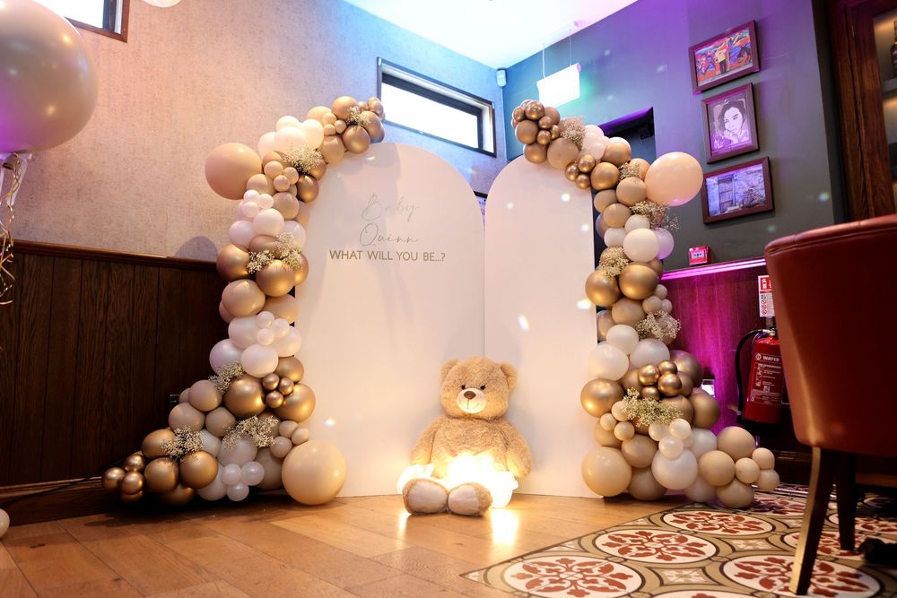 Gender reveal party setup with balloon arch, gold and white balloons, and large teddy bear centerpiece.