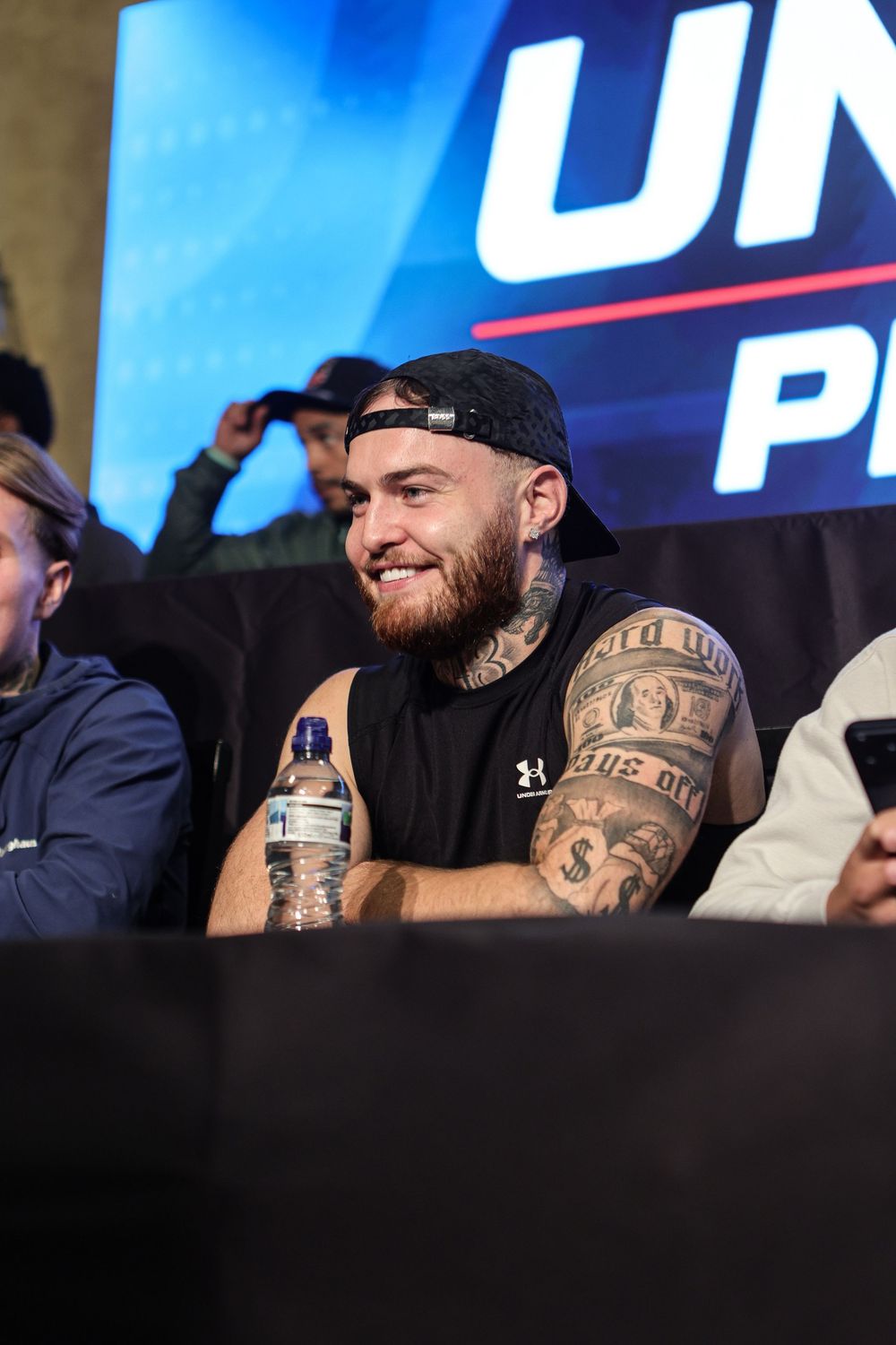 Tattooed man with backward cap smiles at UFC press conference event, wearing black sleeveless shirt.