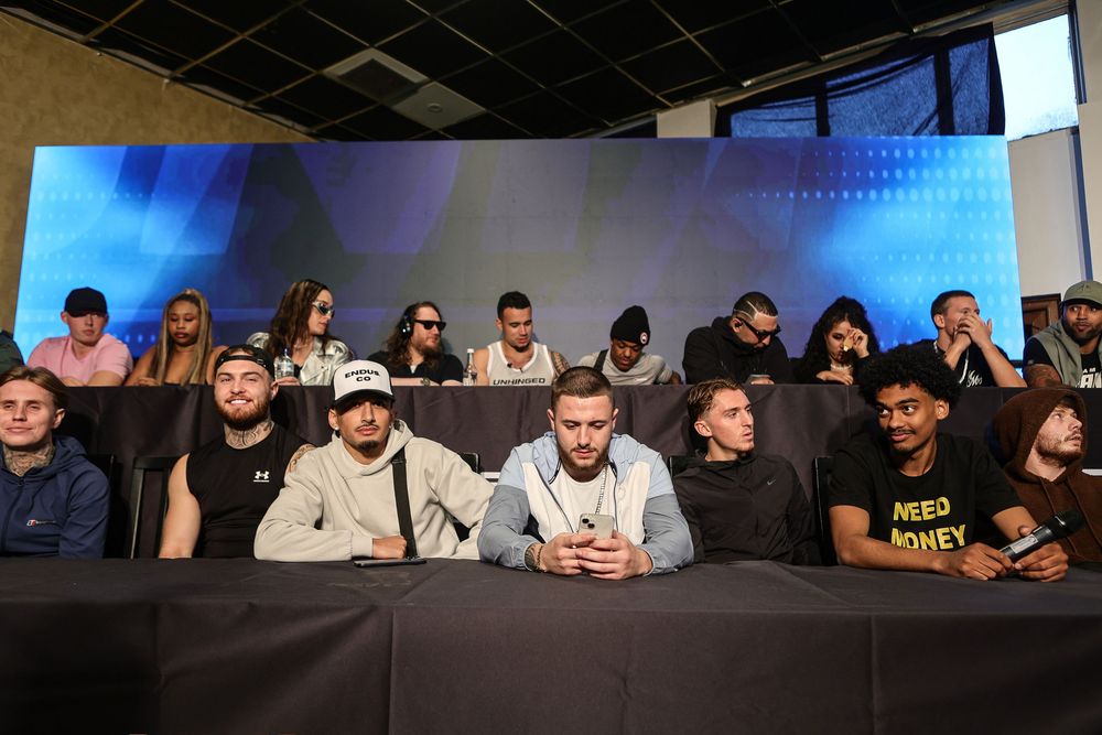 Group of young adults seated in rows at an indoor event or press conference, with a large blue screen backdrop.