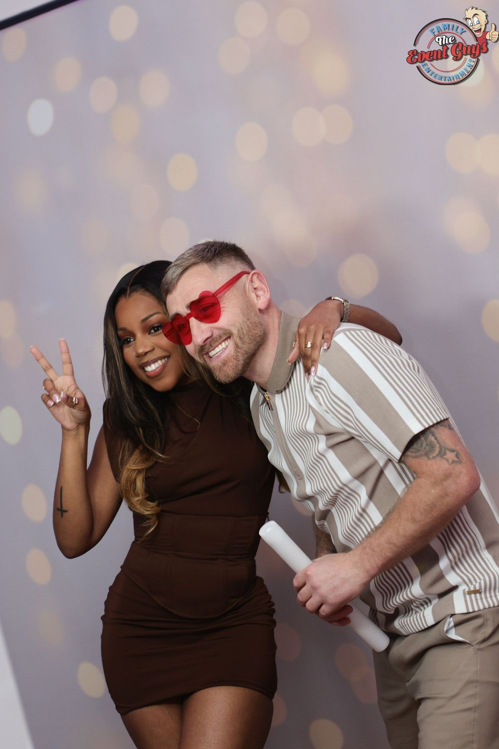 Couple posing at photo booth, woman in brown dress making peace sign, man in heart sunglasses.