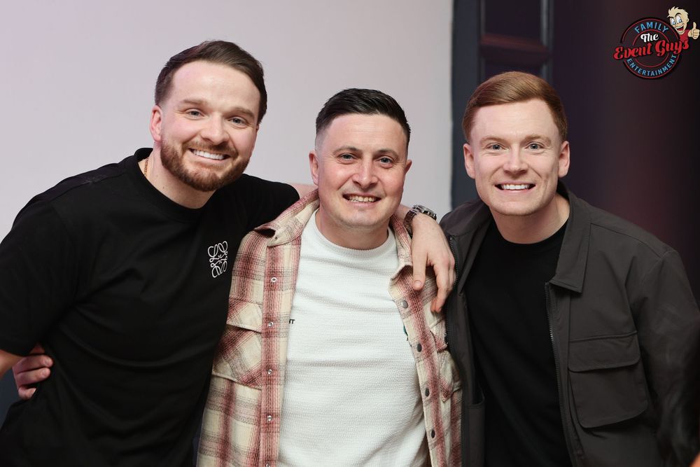 Three men smiling and posing together with arms around each other at an indoor event or venue.