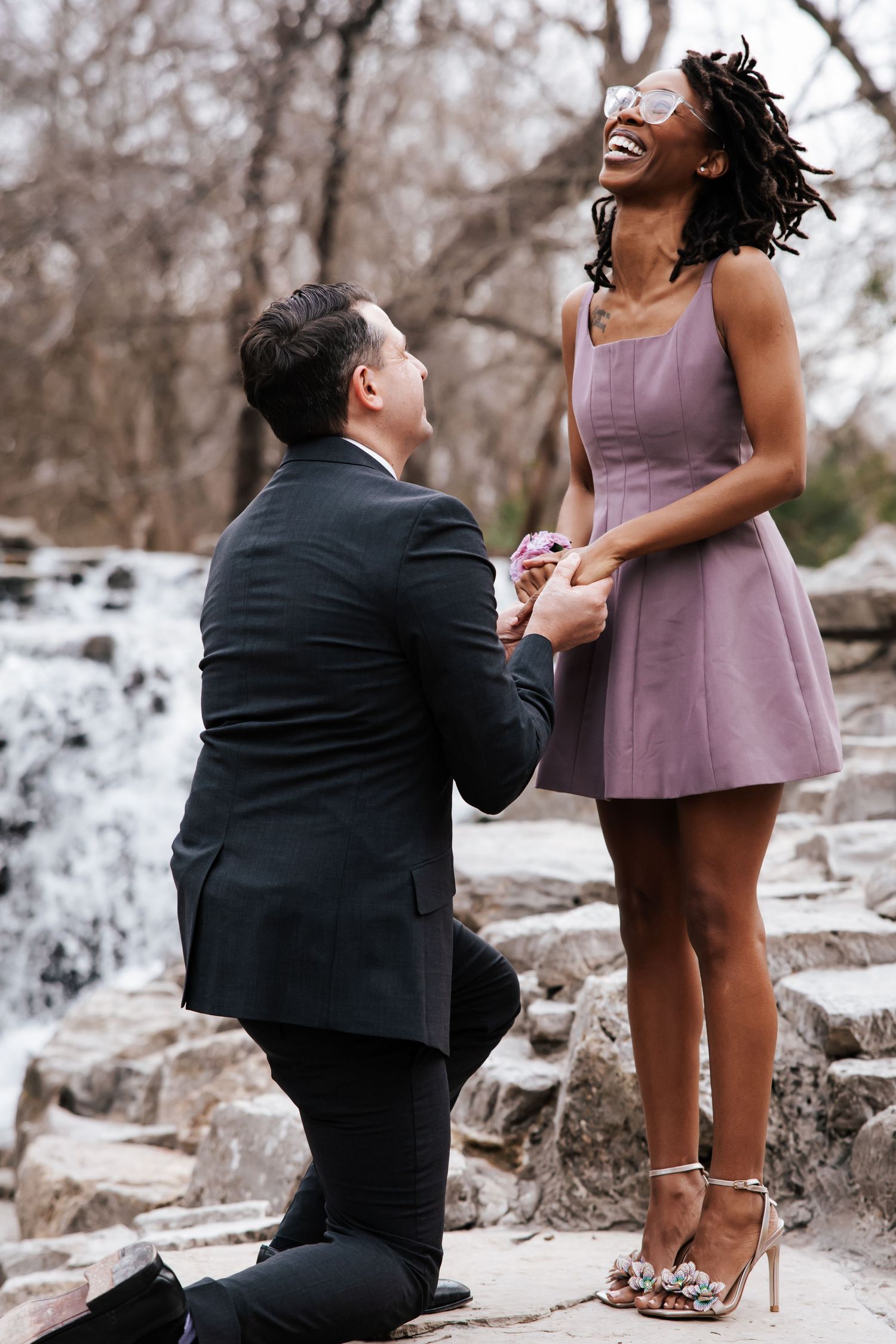 A man proposes on one knee to a joyful woman in a purple dress near a waterfall.