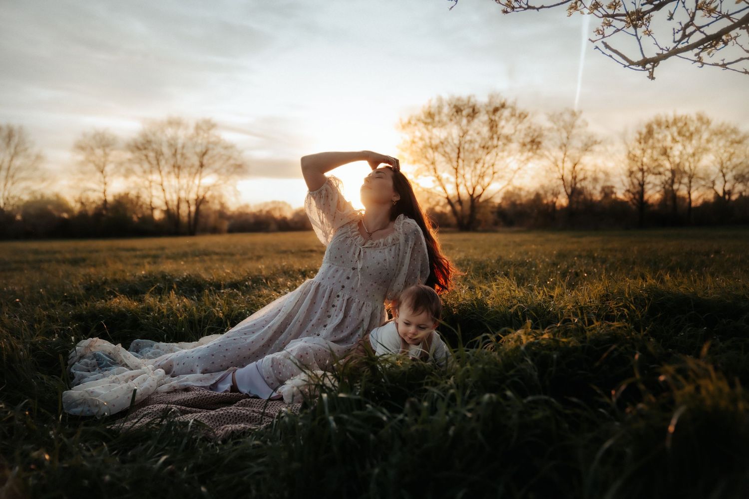 Woman in white dress sitting in a golden field at sunset with a child lying beside her.