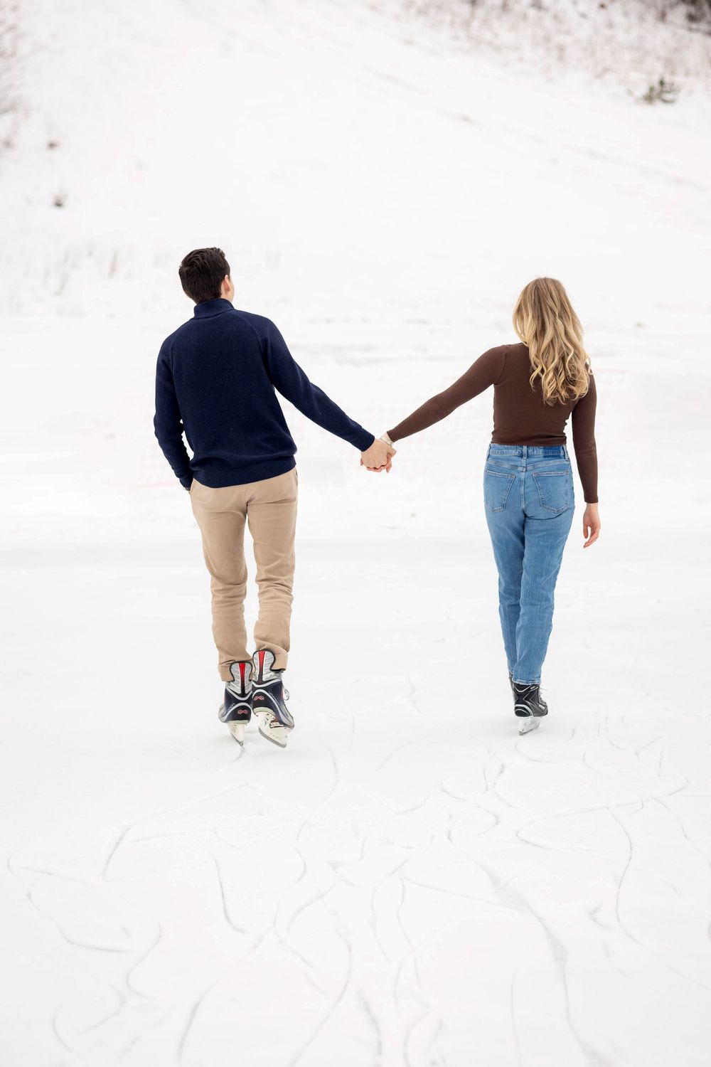 Couple holding hands ice skating on a snowy frozen lake in winter, viewed from behind.