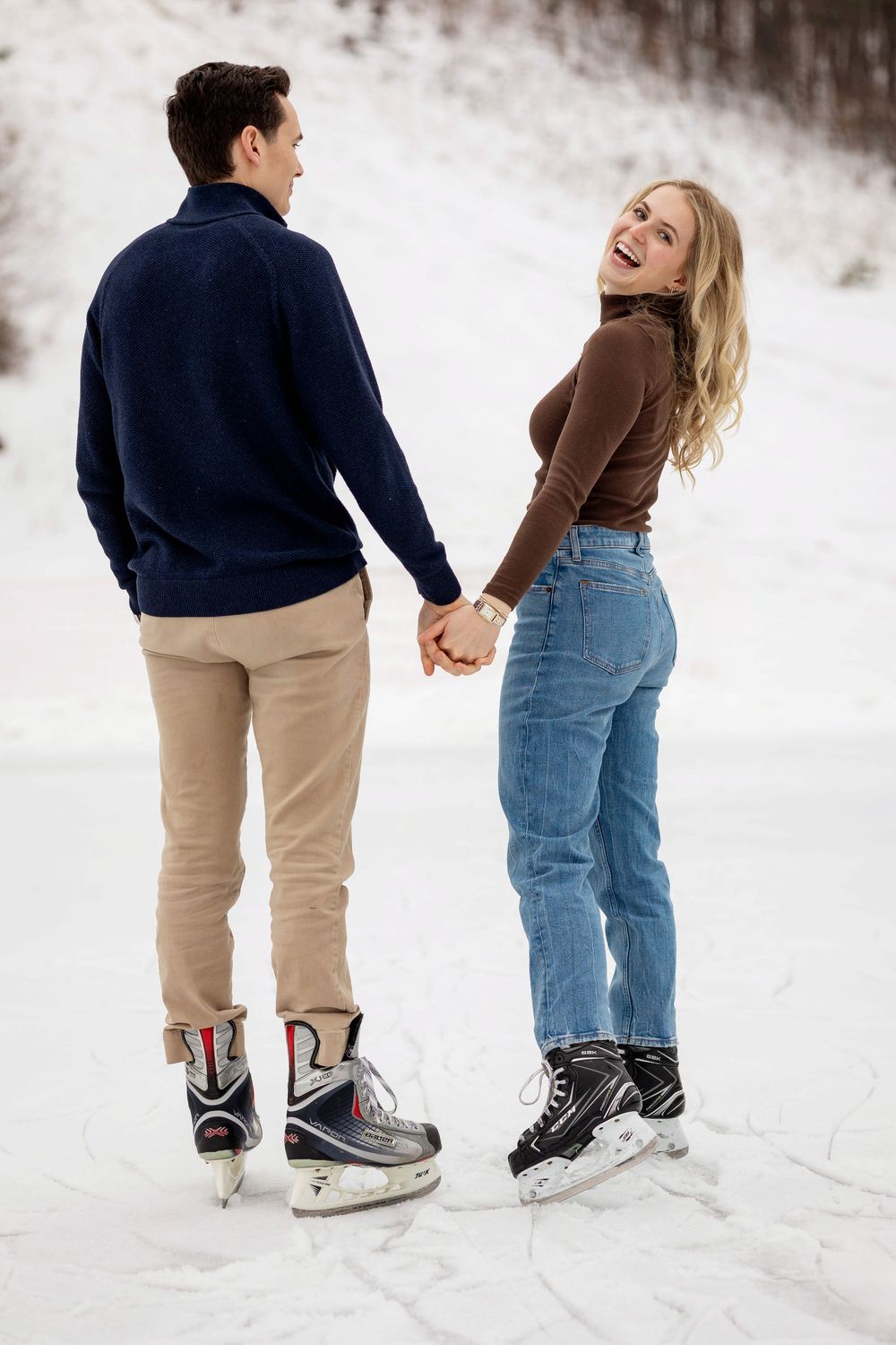 Couple holding hands on outdoor ice rink in winter, woman in jeans smiling back at camera wearing hockey skates.