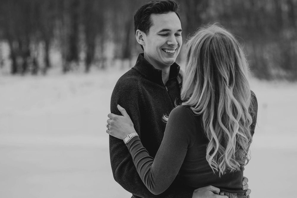 Black and white photo of a smiling couple embracing in a snowy winter landscape during an engagement session.