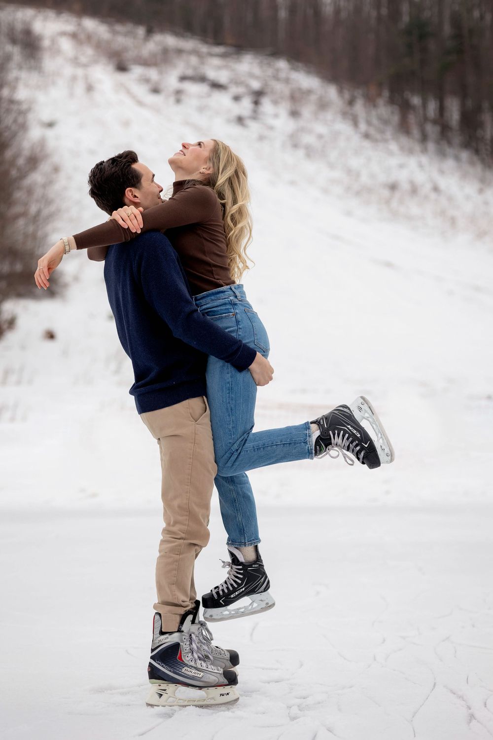 Couple ice skating on frozen pond, man lifting woman in joyful winter embrace, snow-covered hillside background.