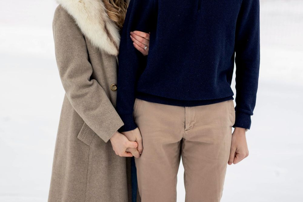 Couple holding hands in winter, wearing a beige coat and navy sweater with tan pants outdoors.