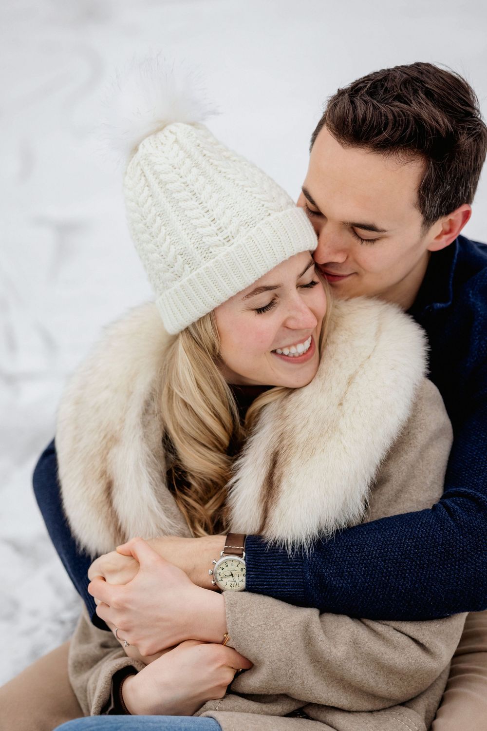 A happy couple embraces in a snowy winter setting, woman in white knit hat and fur coat, man in navy sweater.