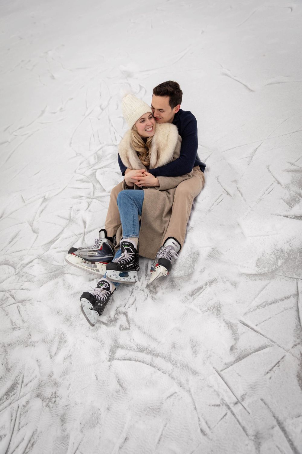 Couple sitting on ice rink in winter, wearing ice skates, sharing a romantic embrace outdoors.