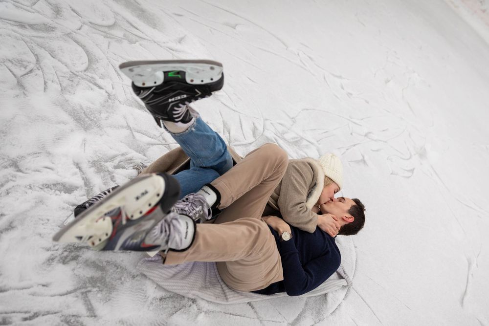 Person laying on ice rink wearing ice skates, lying on back on white ice surface.