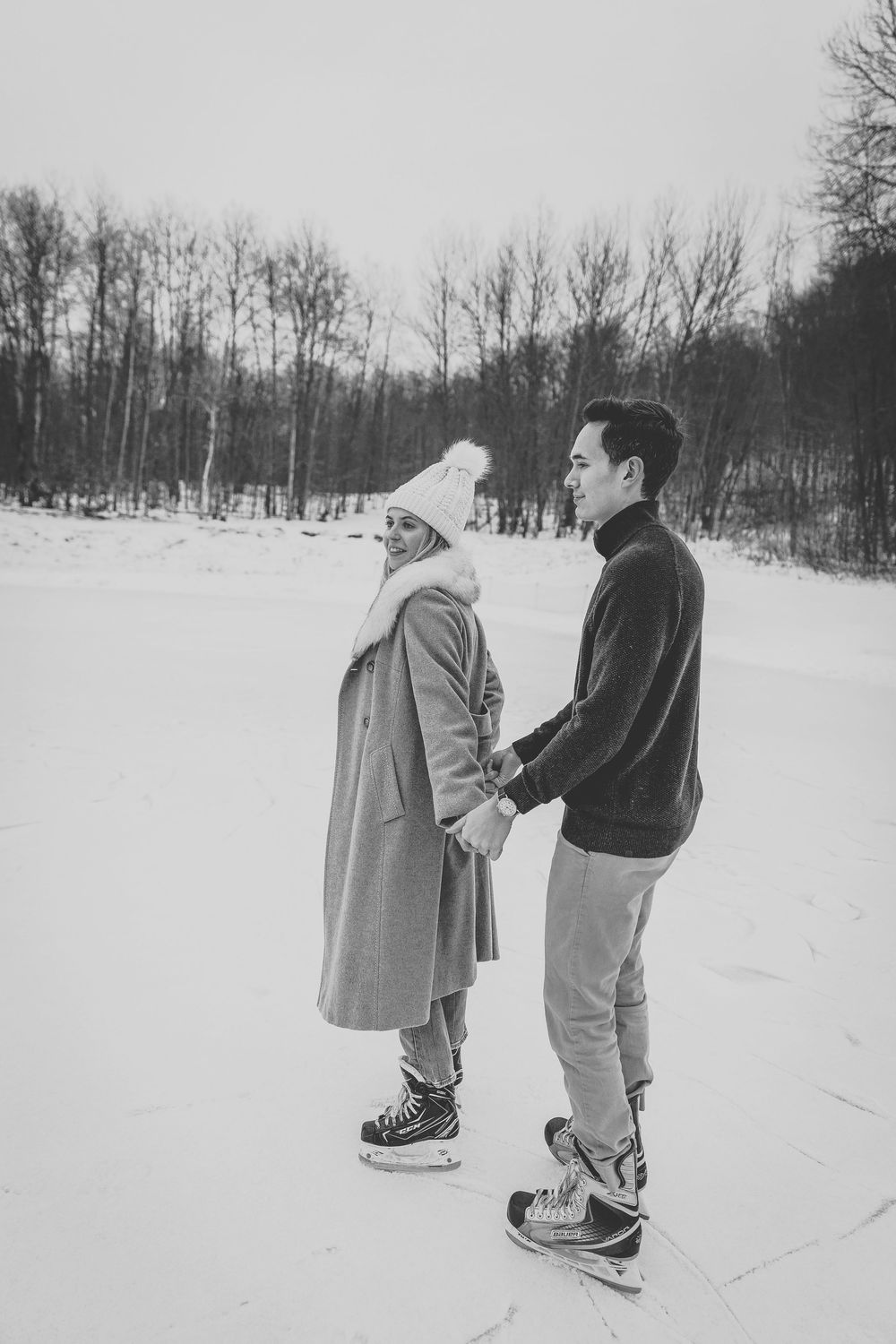 Black and white photo of a couple holding hands while ice skating on a frozen outdoor rink surrounded by snowy trees.