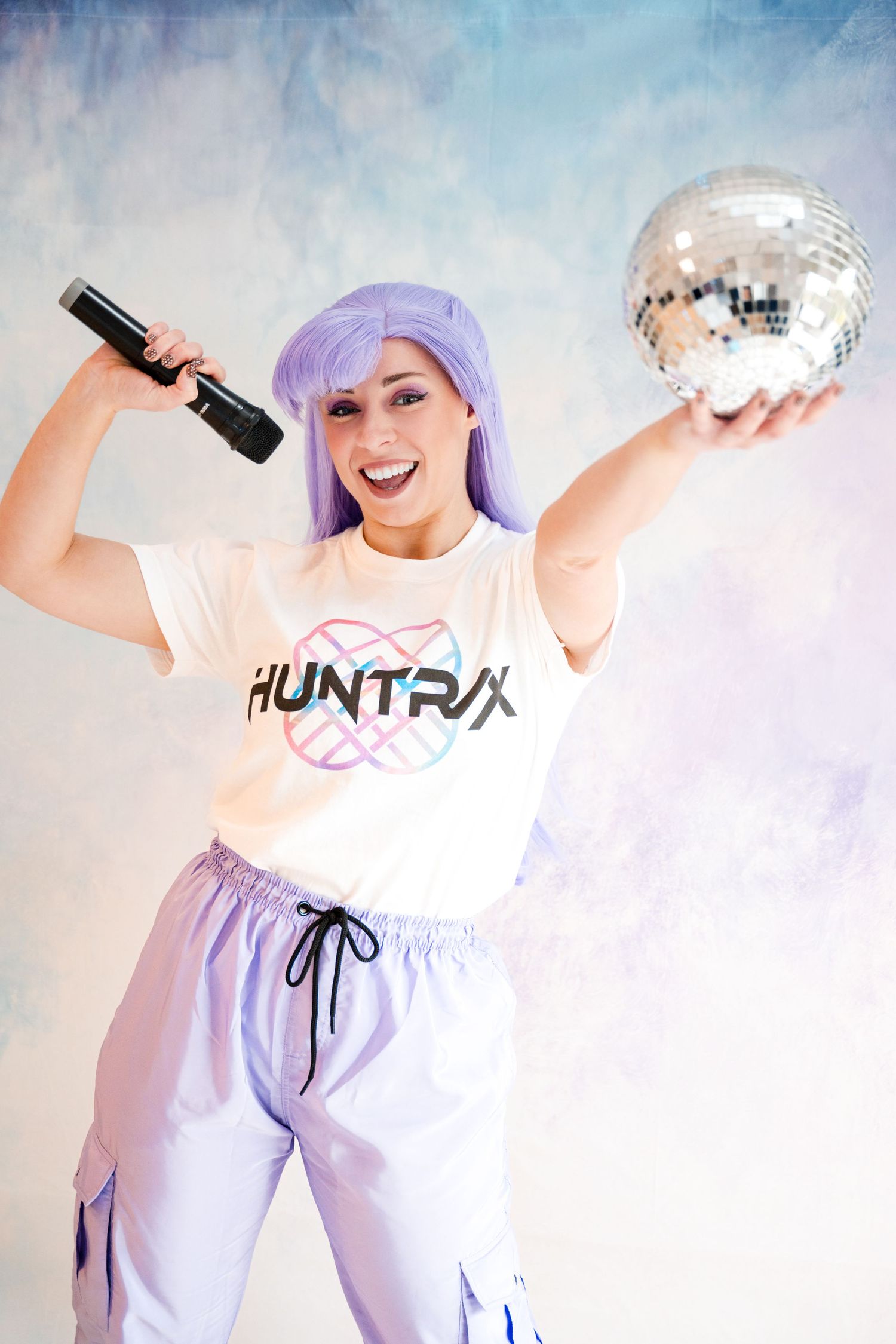 Woman in purple wig holds microphone and disco ball wearing Huntrix branded outfit.