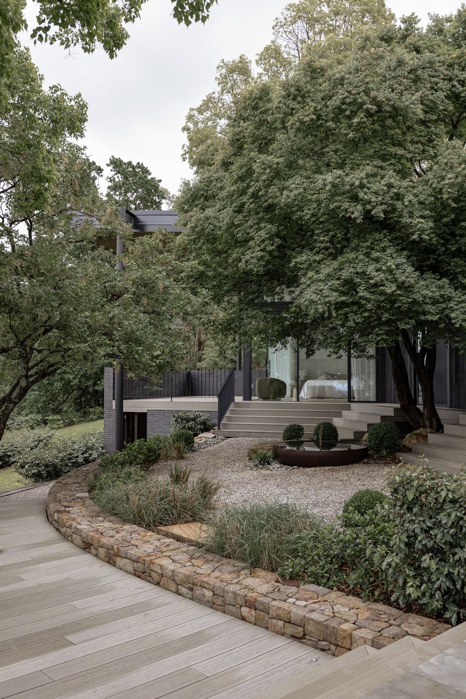 Curved stone retaining wall borders a gravel garden with manicured shrubs leading to a dark modern home with timber deck.
