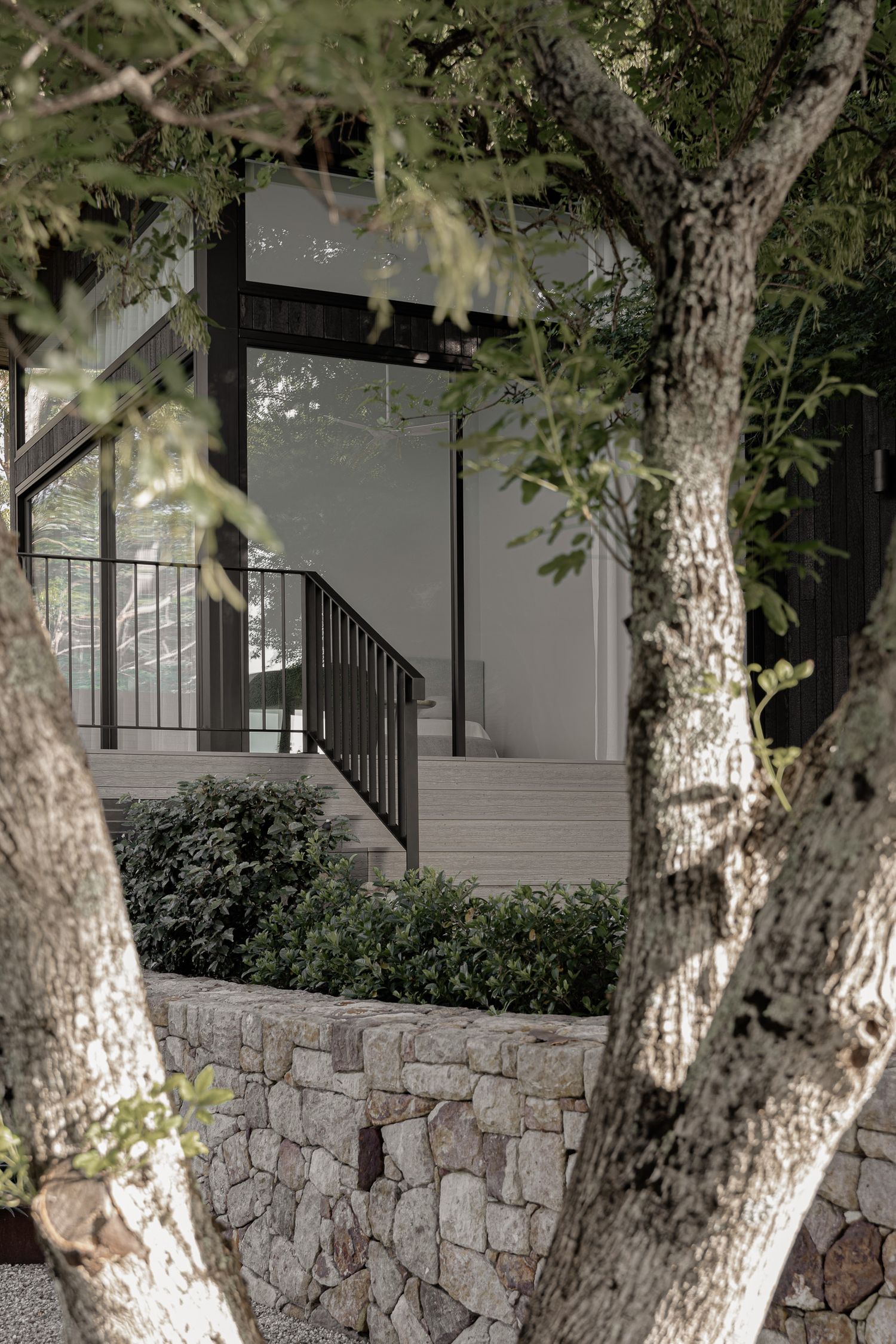 Stone retaining wall with mature trees framing a modern home entrance with black steel staircase railing.