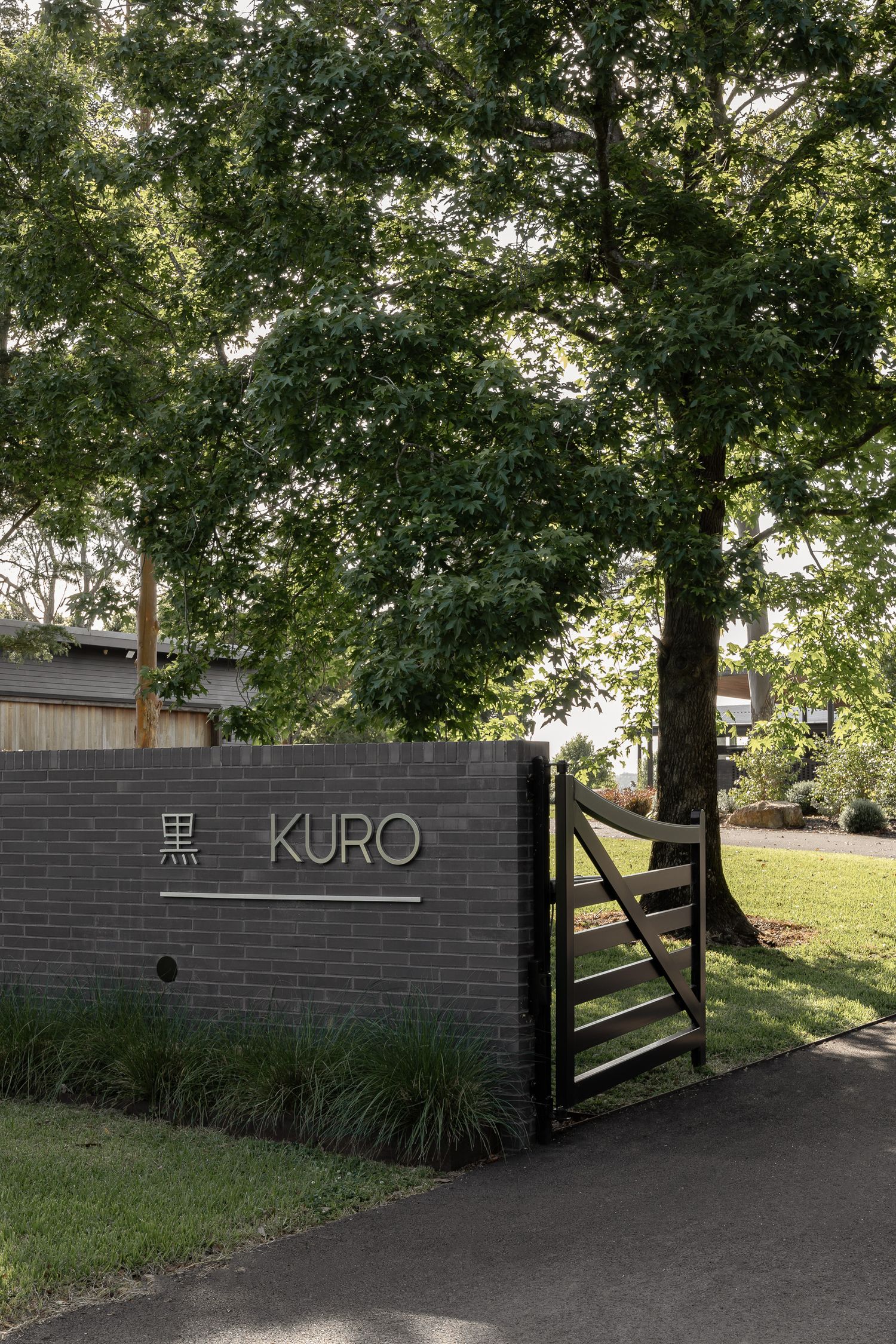 Dark brick wall with 'KURO' sign and wooden gate surrounded by lush green trees at a modern property entrance.