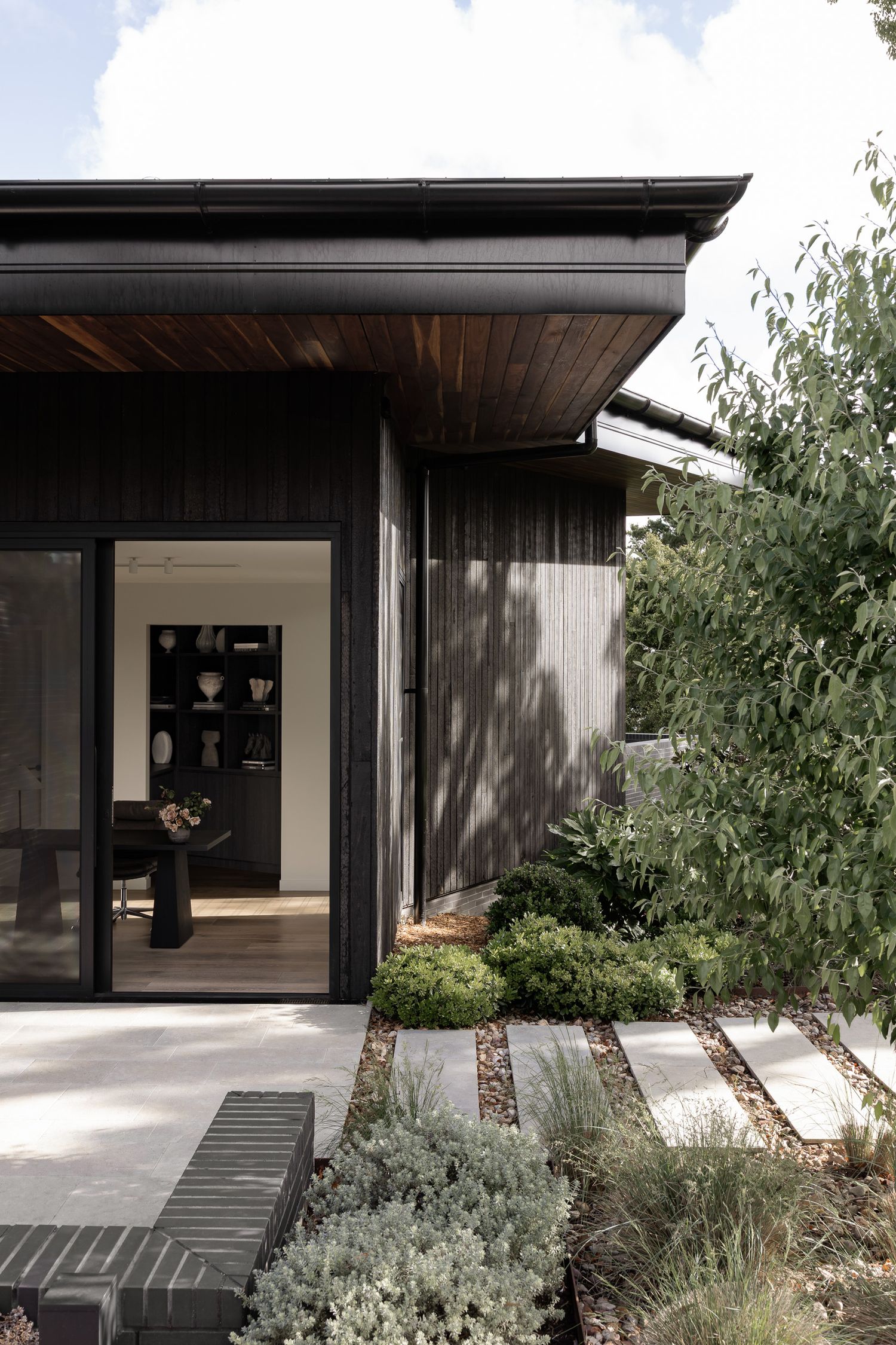 Black timber clad contemporary home with sliding glass doors opening to a stone paved garden with ornamental shrubs.