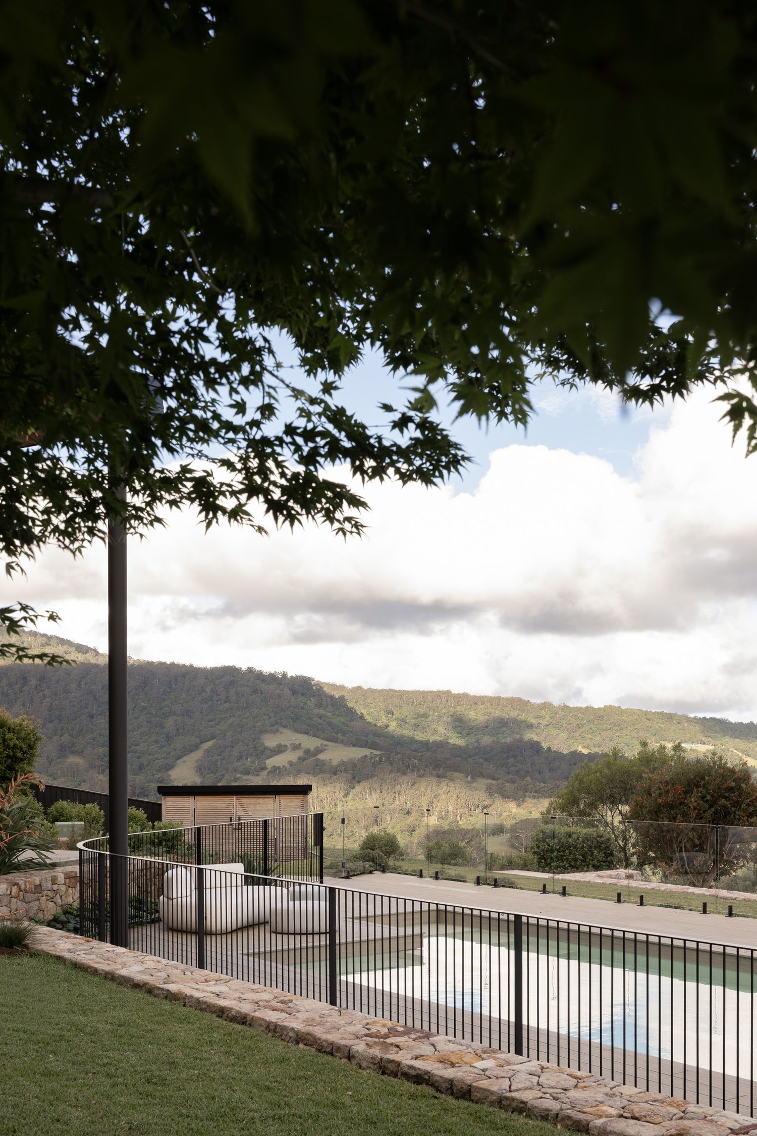 Fenced pool with mountain views framed by Japanese maple tree branches in a lush green landscape.