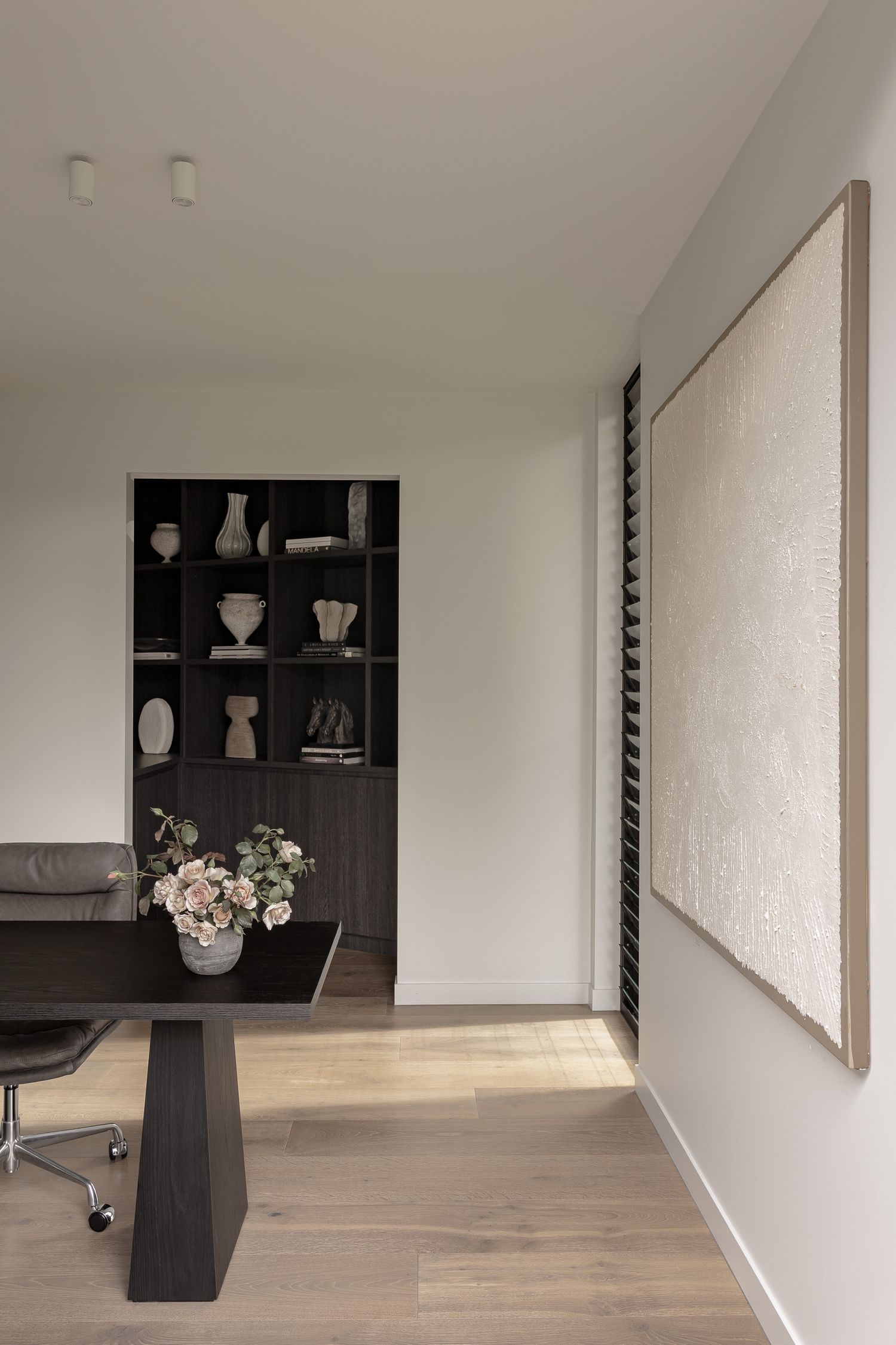 Minimalist home office with dark wood desk, black shelving unit displaying ceramic vases, and large neutral artwork on white wall.