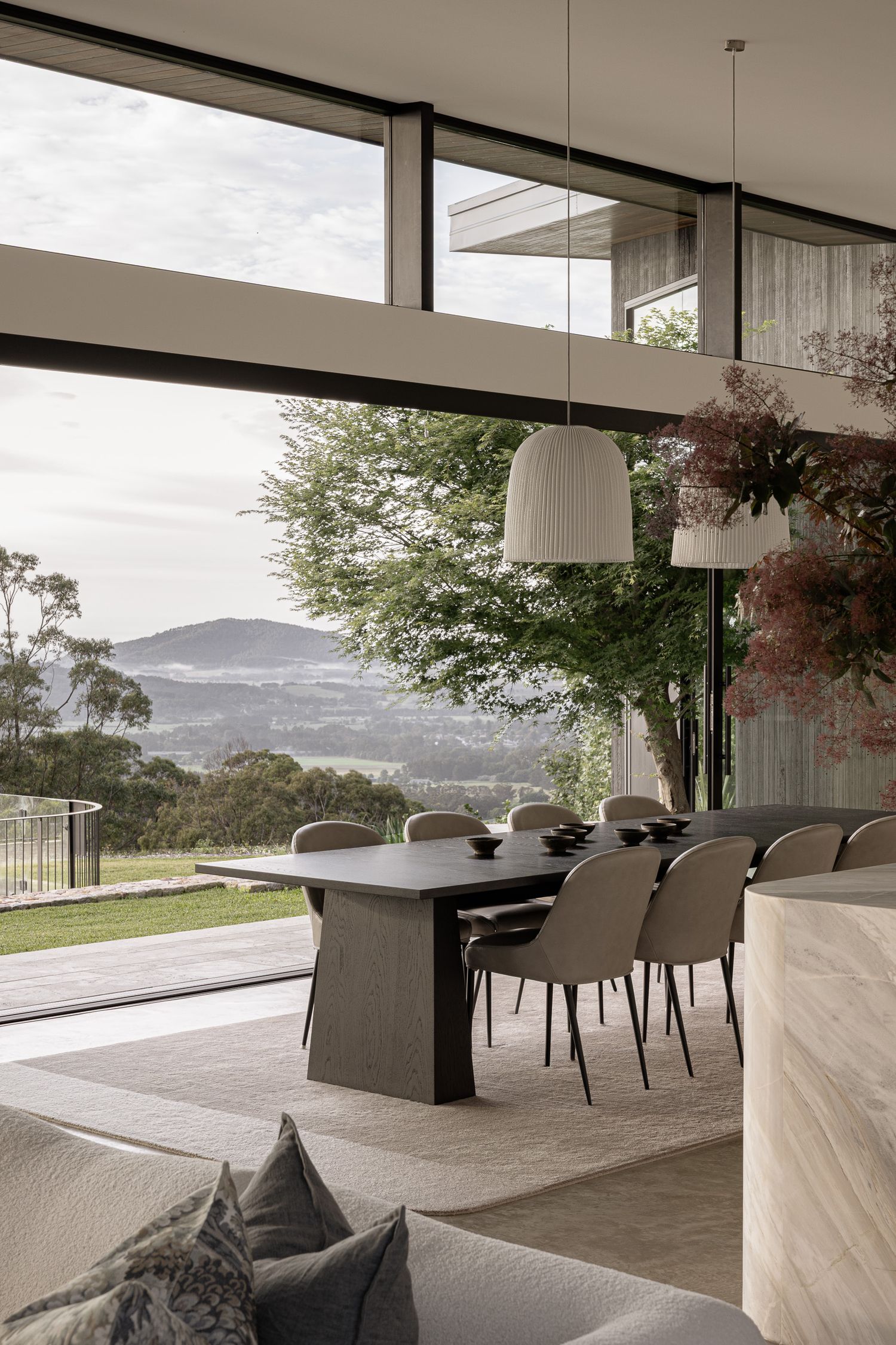 Modern dining room with floor-to-ceiling windows overlooking lush green hills, featuring a dark table, beige chairs, and pendant lights.