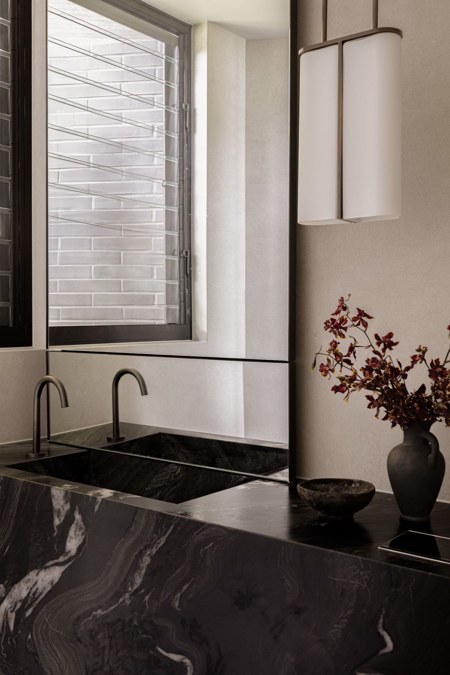 Black marble bathroom vanity with dual sinks, matte faucets, and a vase of dried florals beside a louvered window.