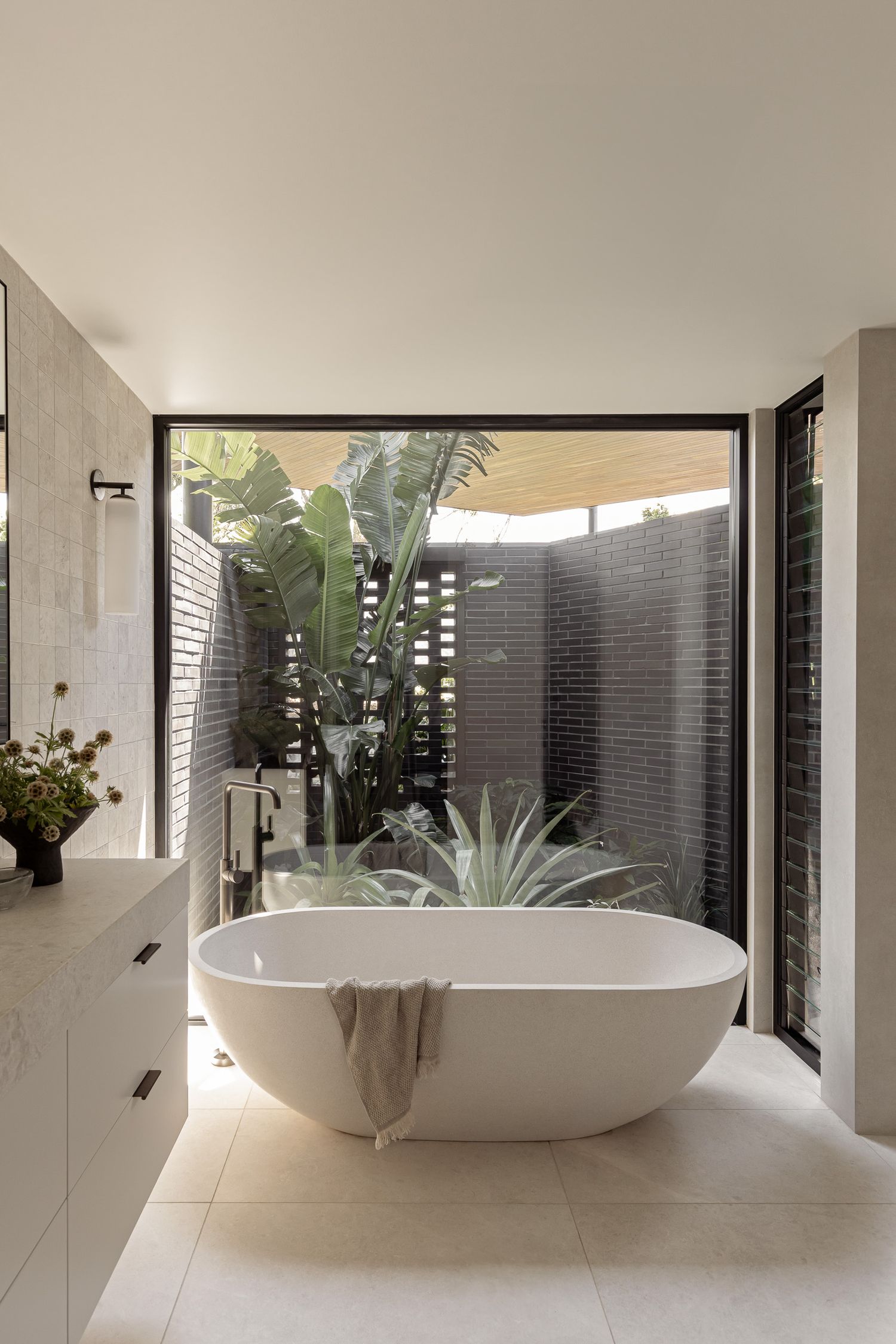 Luxurious freestanding white soaking tub beside floor-to-ceiling windows overlooking a lush tropical garden courtyard.