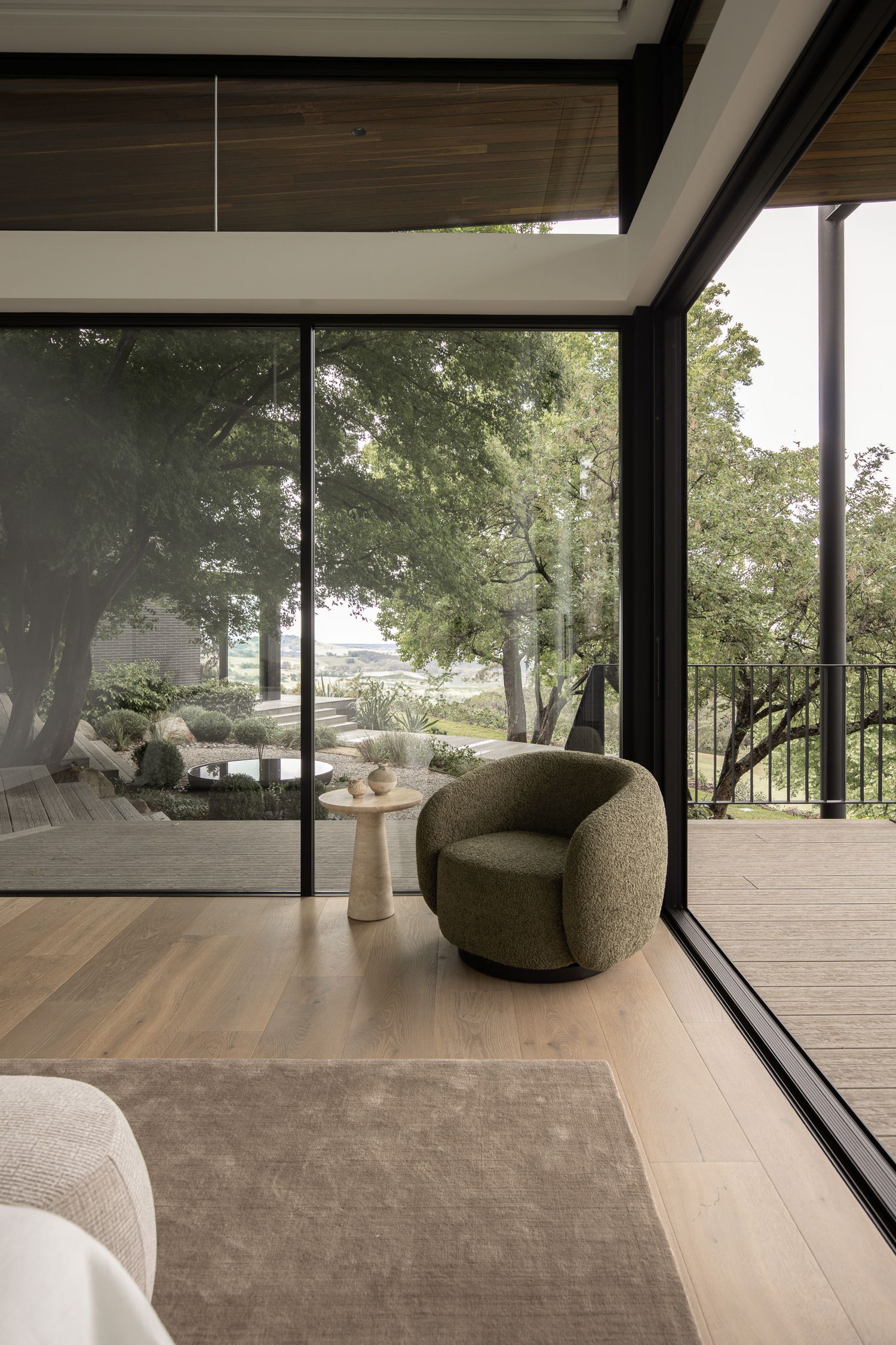 Modern living room with floor-to-ceiling glass walls, green barrel chair, round side table, and lush tree views beyond the deck.