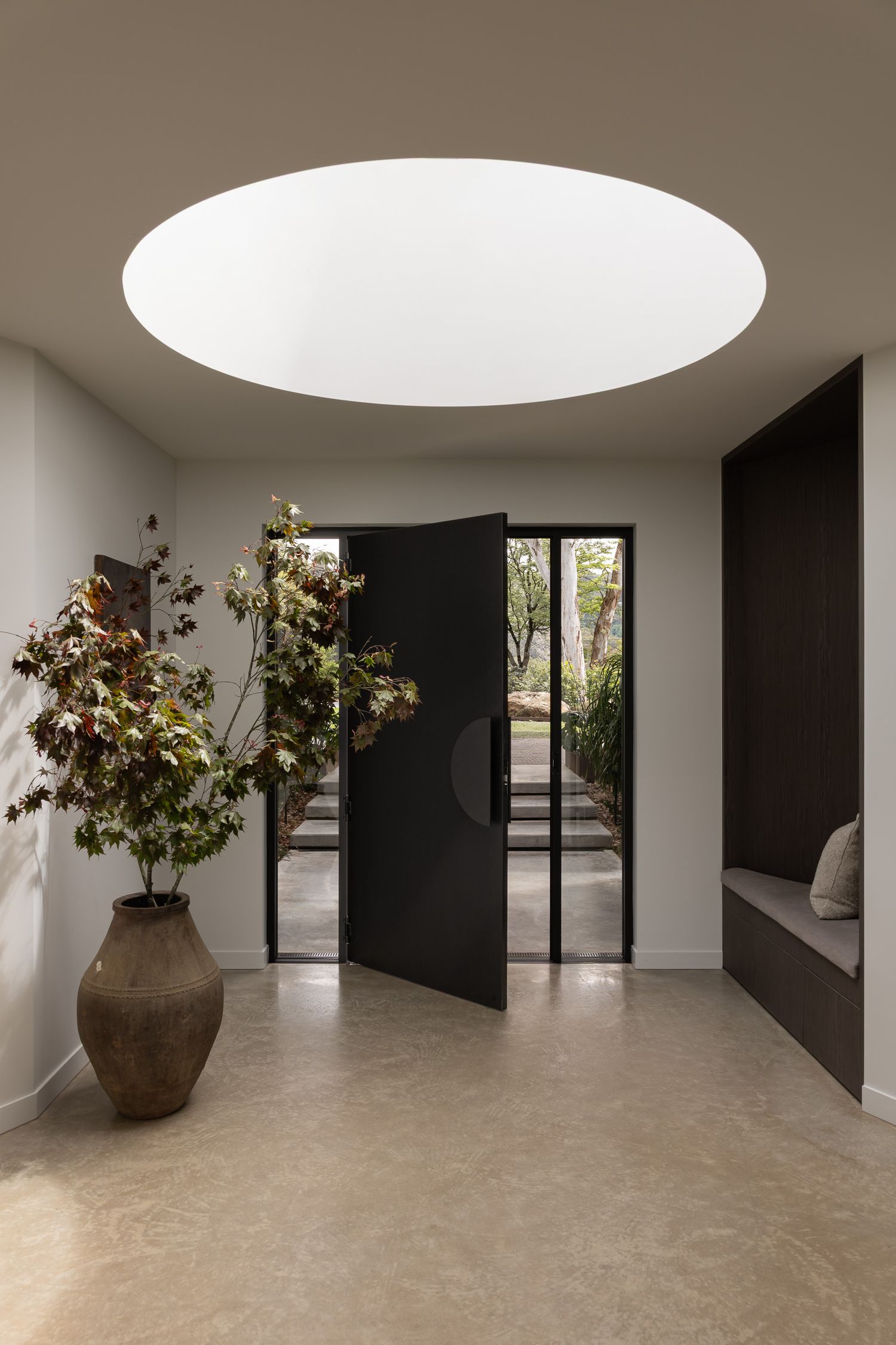 Modern entryway with circular skylight, black pivot door, large ceramic vase with plants, and built-in bench on stone floor.