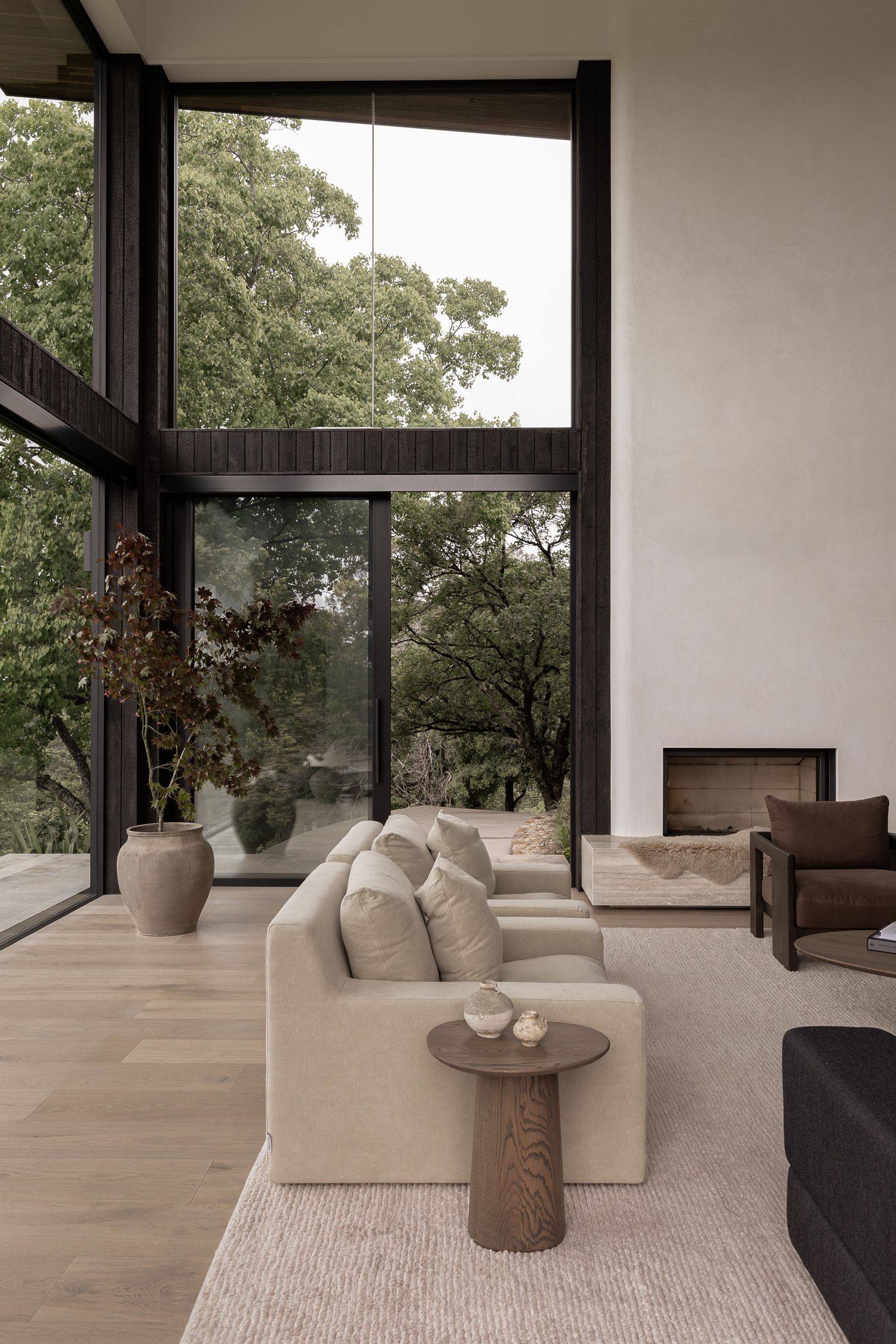Modern minimalist living room with floor-to-ceiling black-framed windows, neutral linen sofa, wood coffee table, and fireplace.