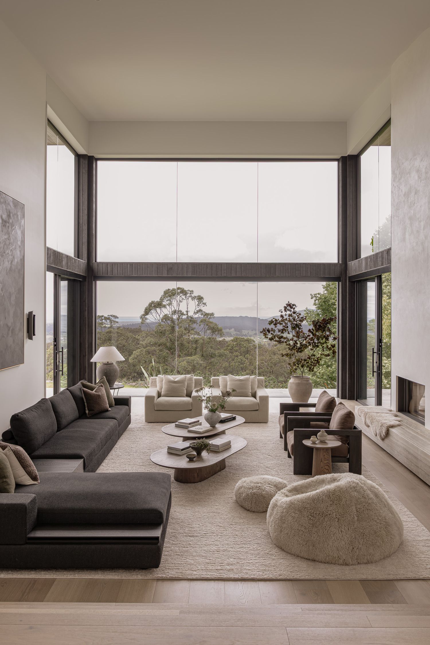 Modern living room with floor-to-ceiling windows, dark sectional sofa, organic coffee table, and panoramic forest views.