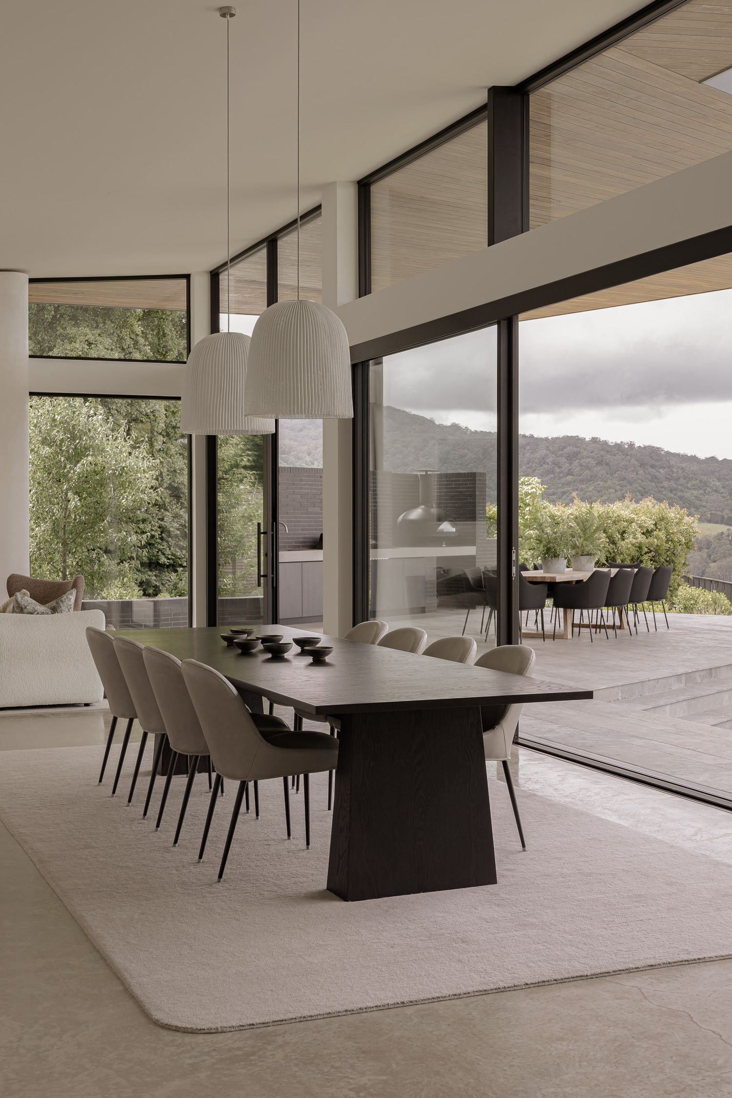 Modern dining room with dark table, beige chairs, pendant light, and floor-to-ceiling windows overlooking a lush hillside.