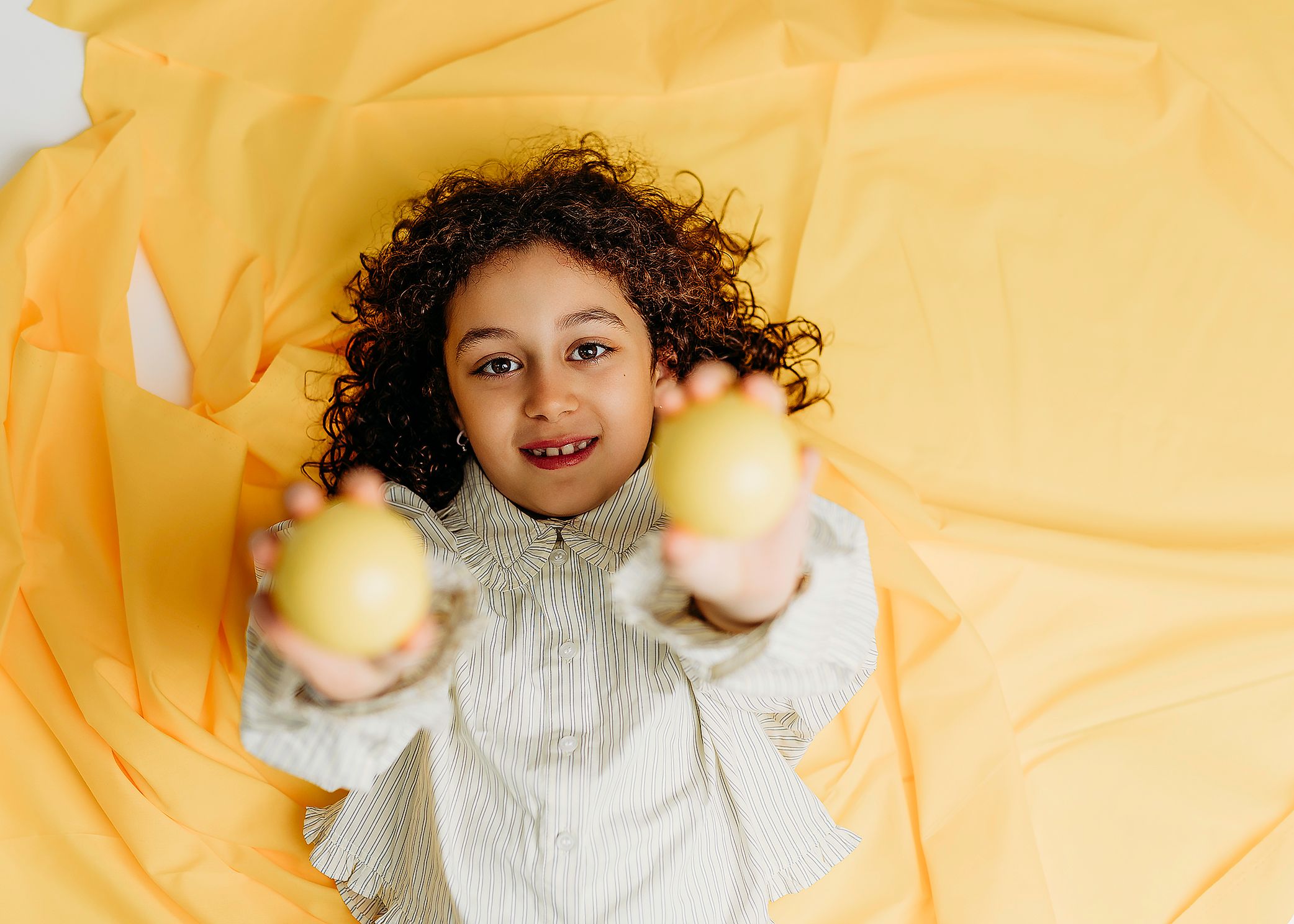Curly-haired child lying on yellow fabric, smiling and holding two yellow balls toward the camera.