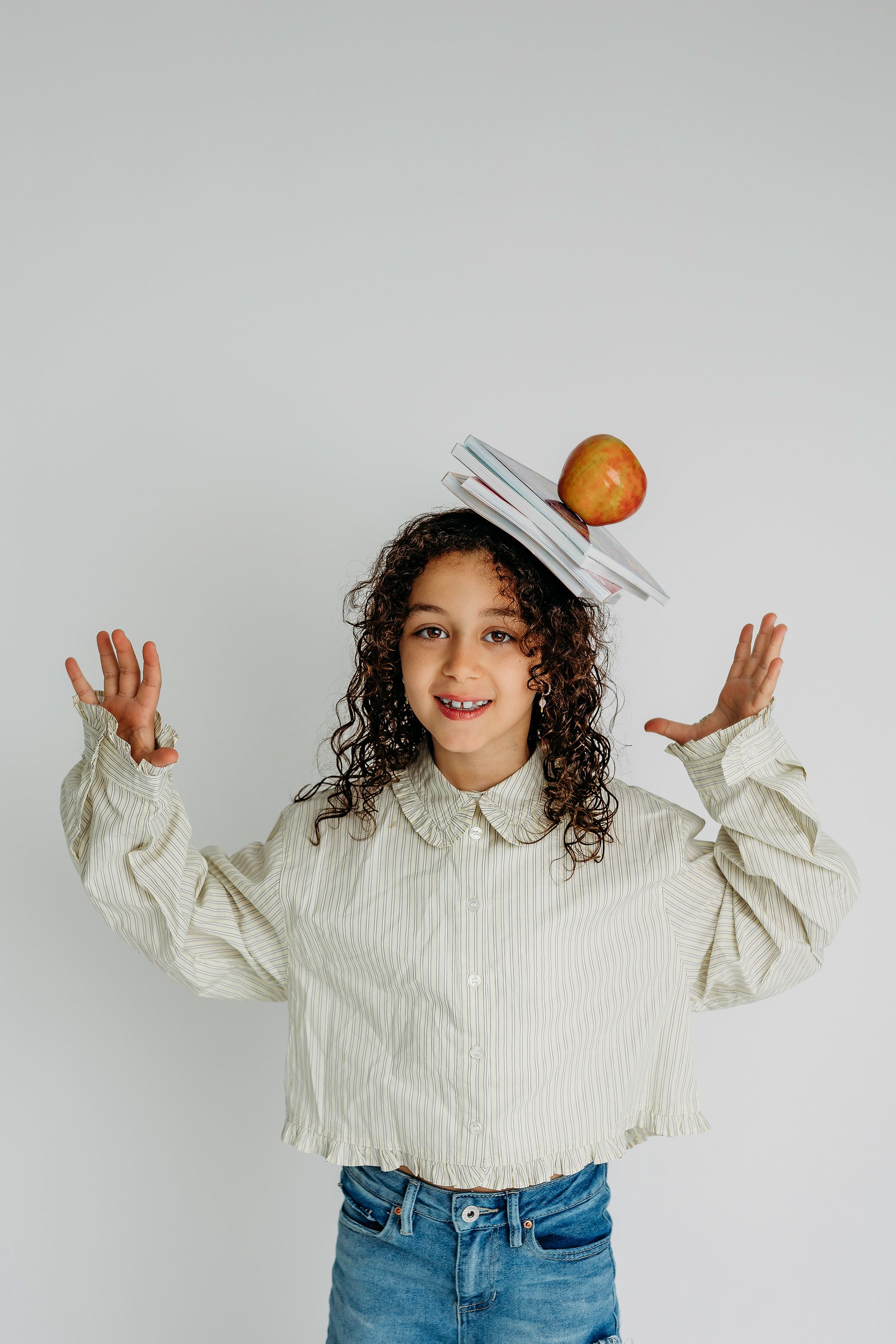 Young girl with curly hair balancing books and an apple on her head, arms raised, wearing a striped shirt and jeans.