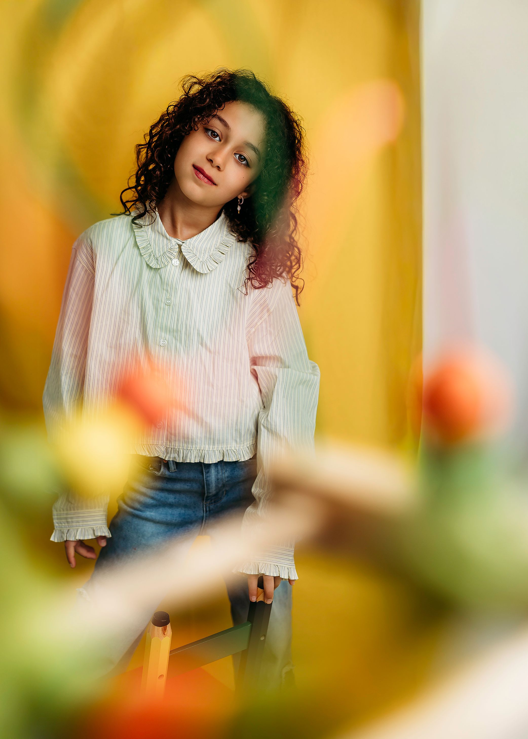 Young woman with curly hair in white blouse and jeans poses thoughtfully among colorful blurred flowers.